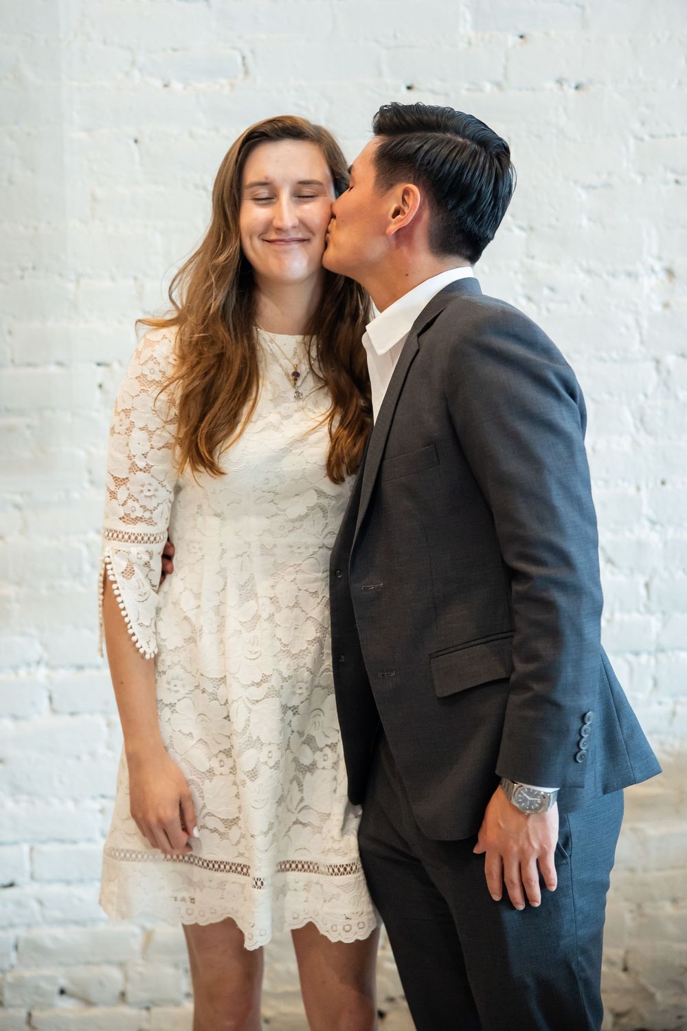 A man in a gray suit kisses a smiling woman in a white lace dress against a white brick wall.