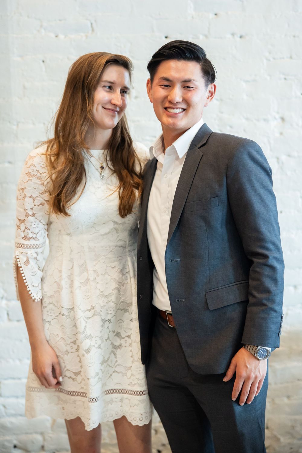 A smiling couple poses together, woman in white lace dress and man in gray suit, against a white brick wall.