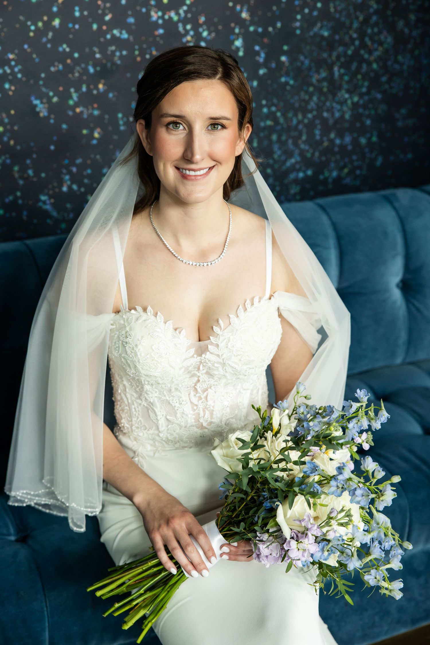 Bride in white gown and veil holds blue and white floral bouquet, smiling softly against a bokeh backdrop.
