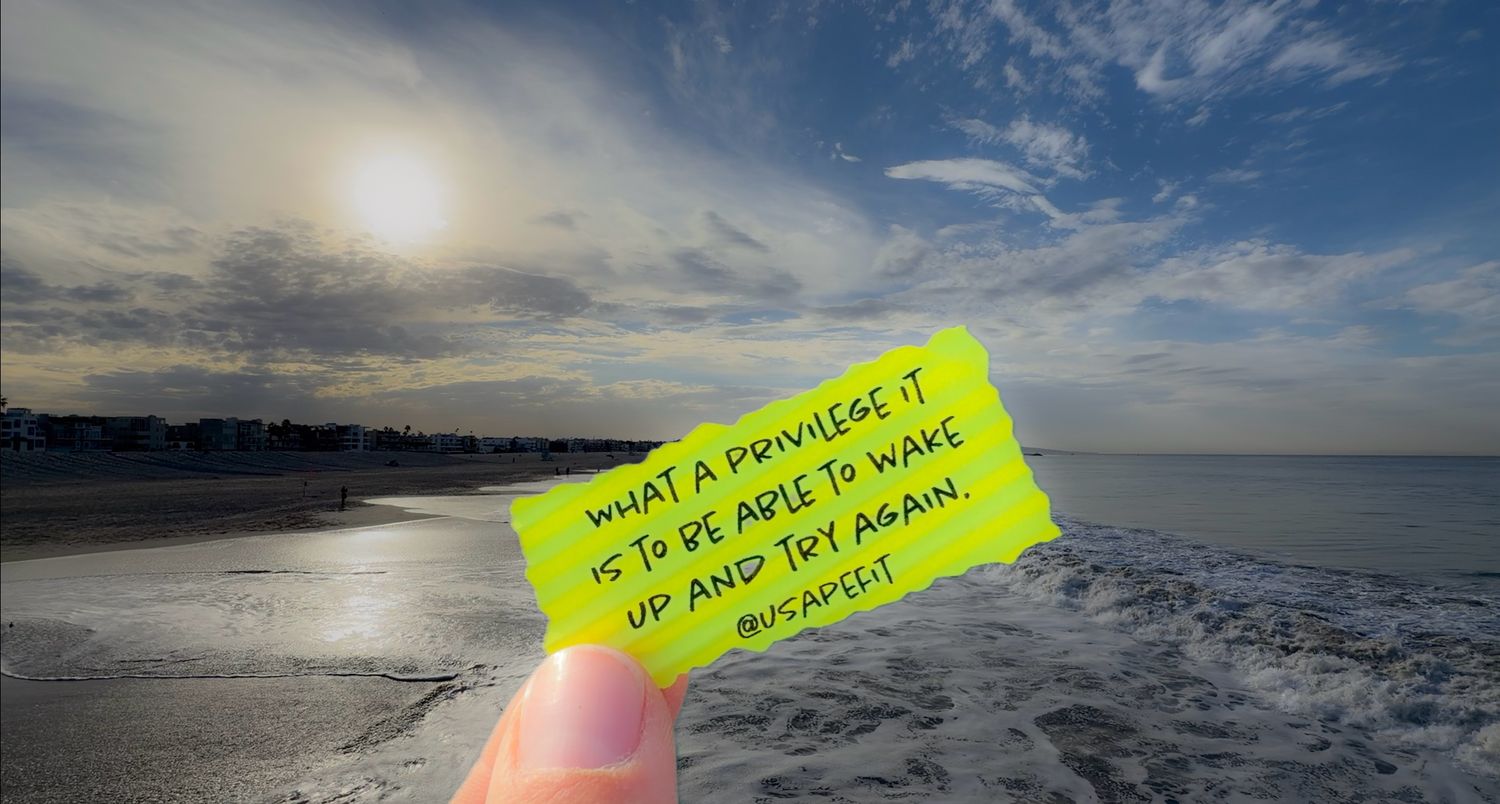 Hand holding a yellow note reading what a privilege it is to be able to wake up and try again over a sunlit beach and ocean waves.