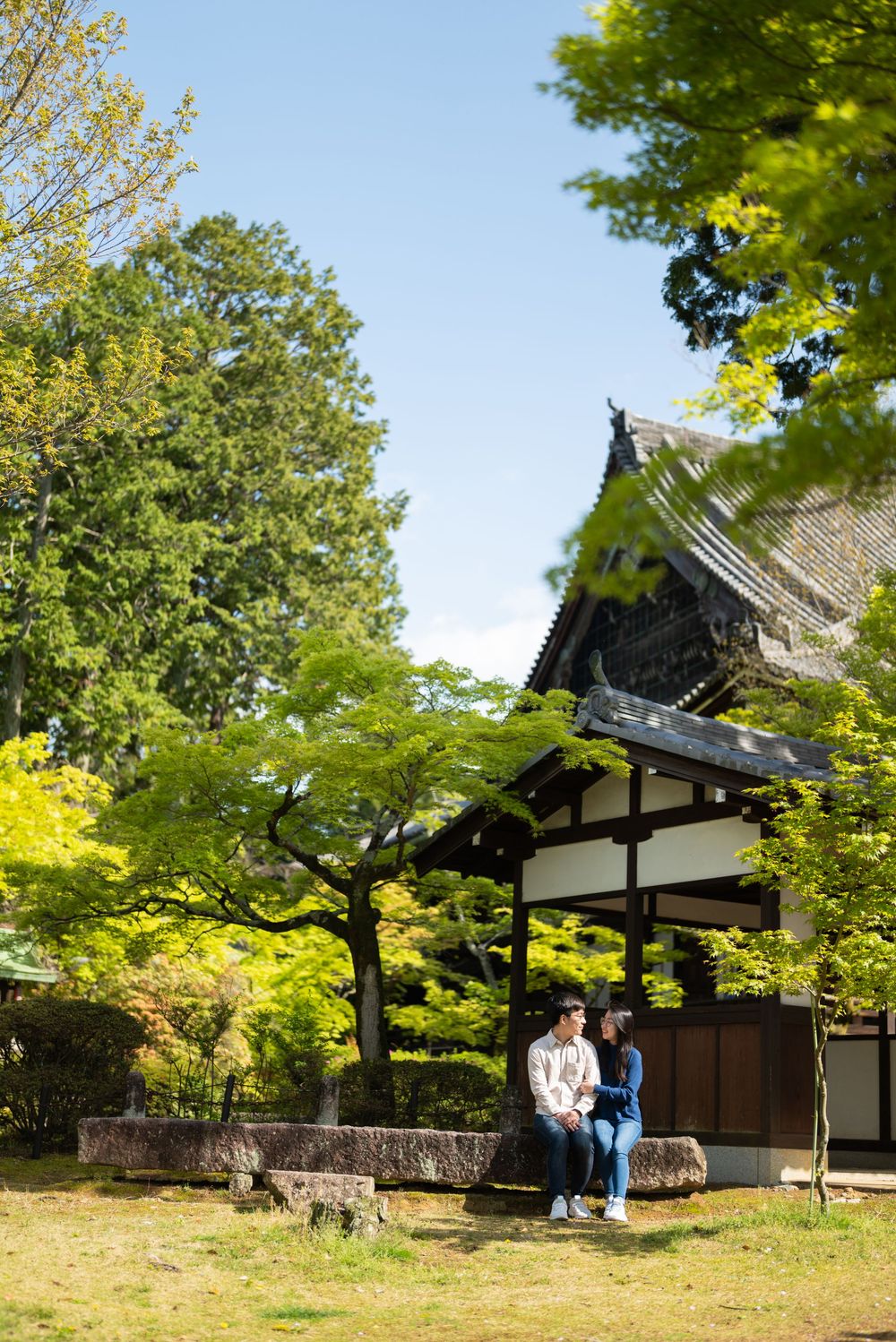 A couple sits together on a stone ledge beside a traditional Japanese temple surrounded by lush green maple trees.