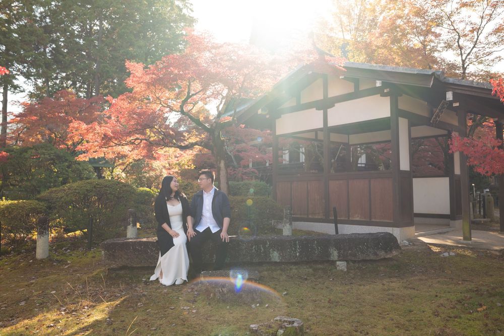 Couple in wedding attire sitting together before a traditional Japanese wooden shrine surrounded by vibrant red autumn maple leaves.
