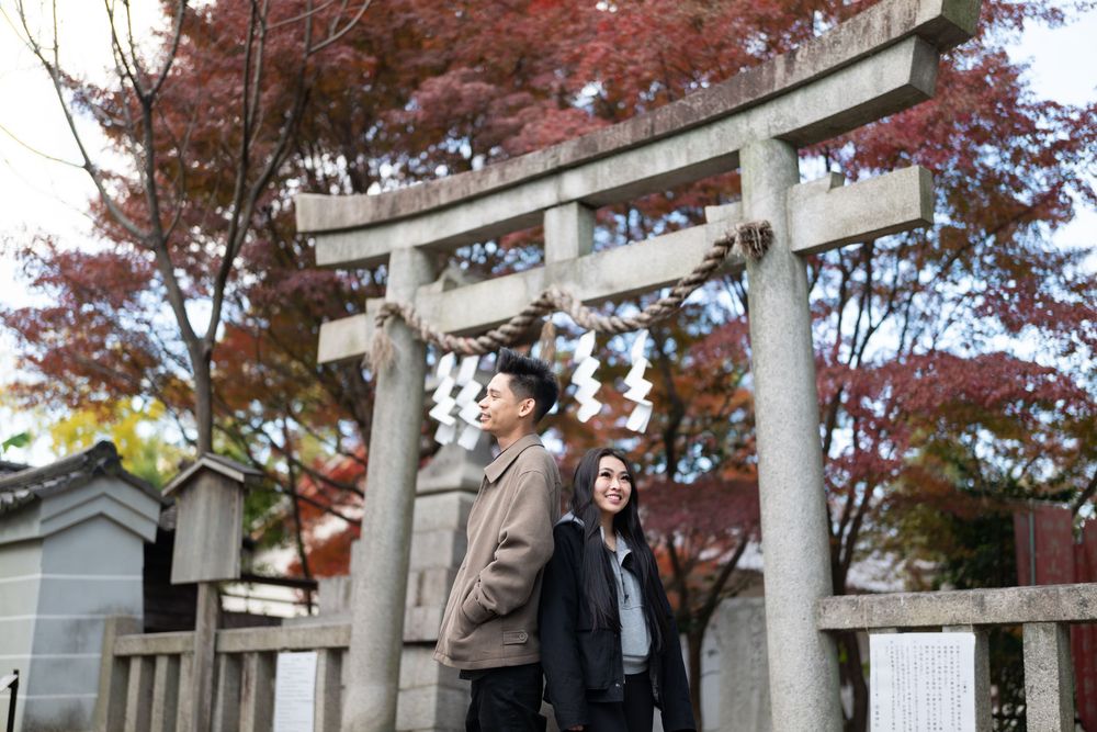 Couple in coats standing beneath a stone torii gate with a shimenawa rope at a Japanese shrine surrounded by red autumn maple leaves.