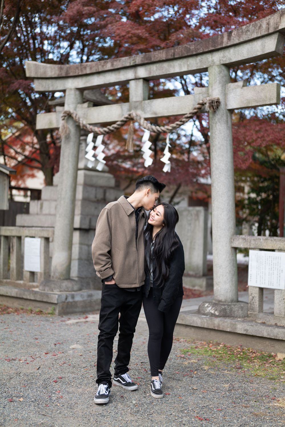 Couple embracing and kissing in front of a stone torii gate with shimenawa rope at a Japanese shrine surrounded by red autumn maple leaves.