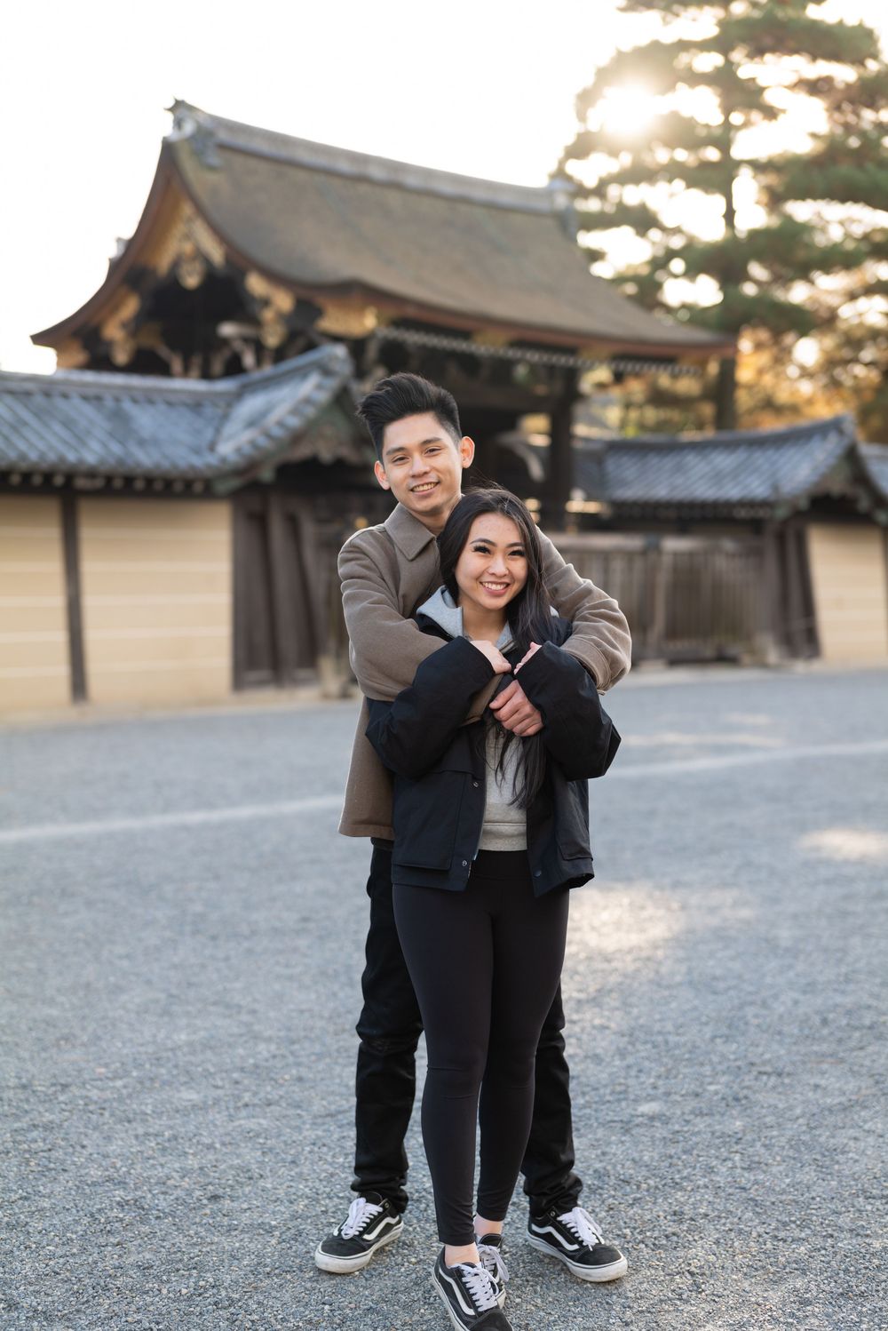 Smiling couple embracing in front of a traditional Japanese temple with a curved tiled roof, gravel courtyard and golden sunset light.