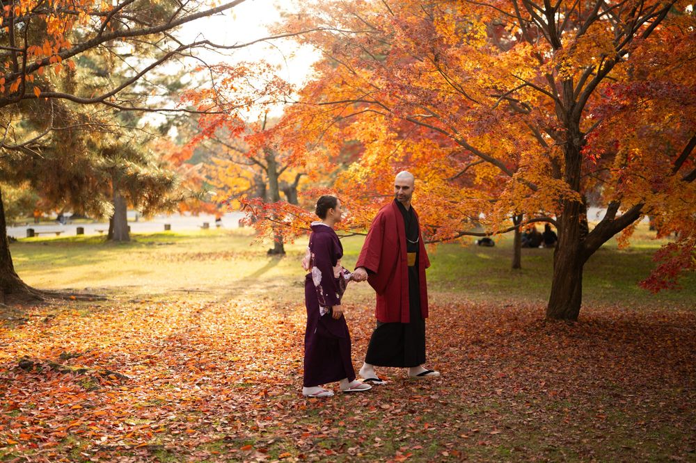Couple in traditional Japanese kimono holding hands and walking through a park covered in fallen red and orange autumn leaves.