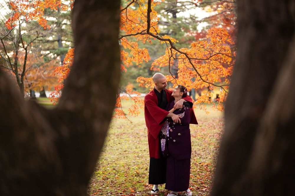 Couple in traditional kimono embracing in a park framed by tree trunks, with vibrant orange autumn foliage glowing in the background.
