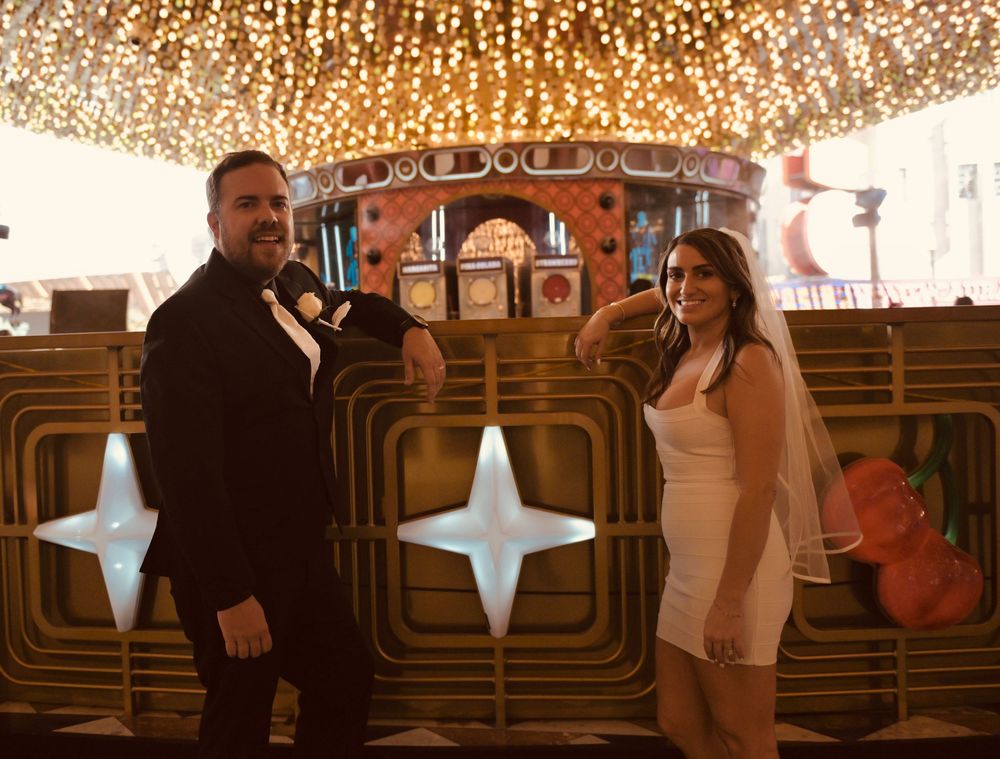 Bride in white mini dress and groom in black tuxedo pose at a glittering Las Vegas venue.