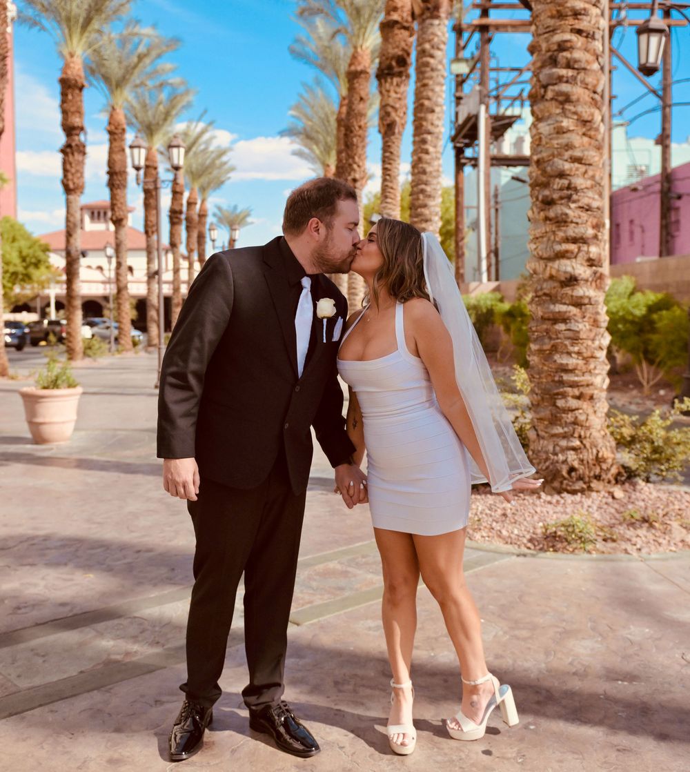 Newlywed couple shares a kiss while holding hands among palm trees in sunny Las Vegas.