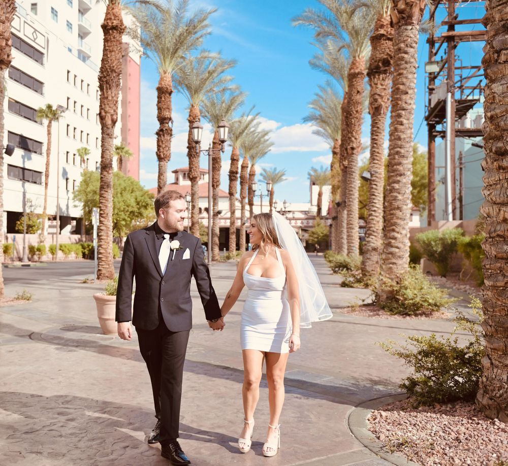 Newlywed couple holding hands walking among palm trees in Las Vegas, bride in white mini dress and groom in black tuxedo.