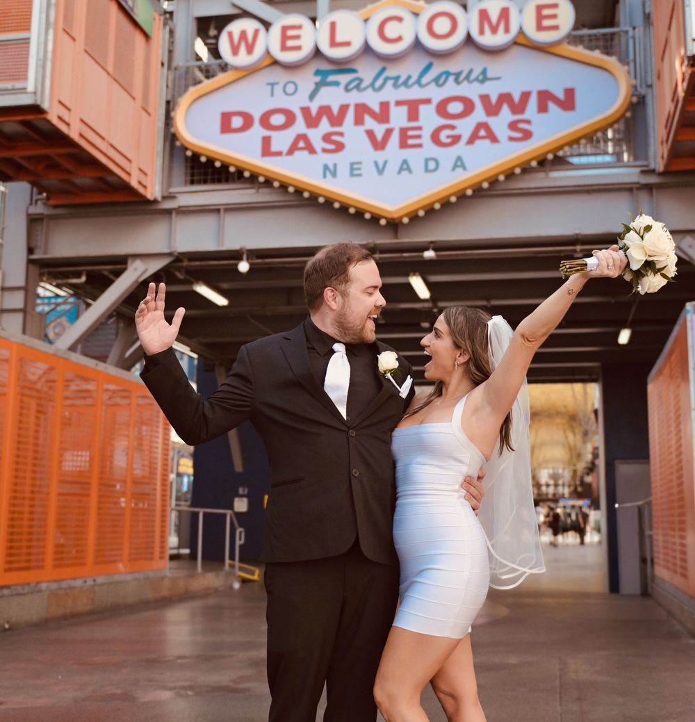 Newlywed couple celebrating in front of Welcome to Downtown Las Vegas sign, bride in white dress holding bouquet.