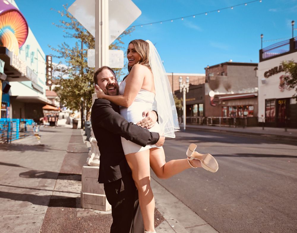 Groom lifting bride in wedding attire on a sunny street, celebrating their marriage outdoors.