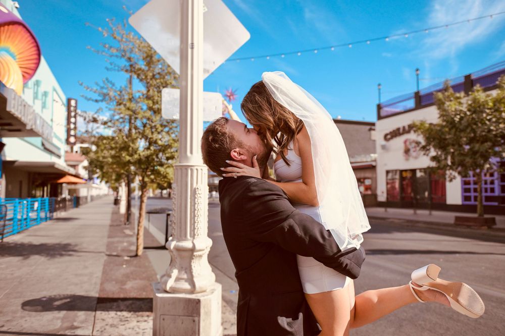 Bride and groom sharing a romantic kiss by a street pole on their wedding day under a bright blue sky.