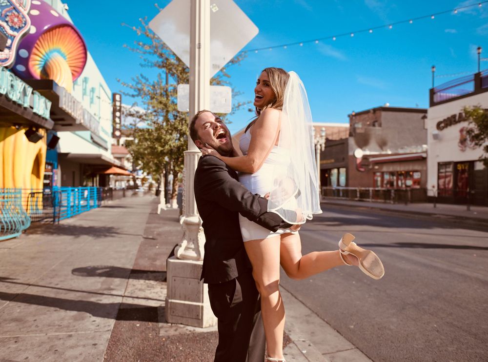 Joyful groom lifting bride in wedding attire on a sunny Las Vegas street.
