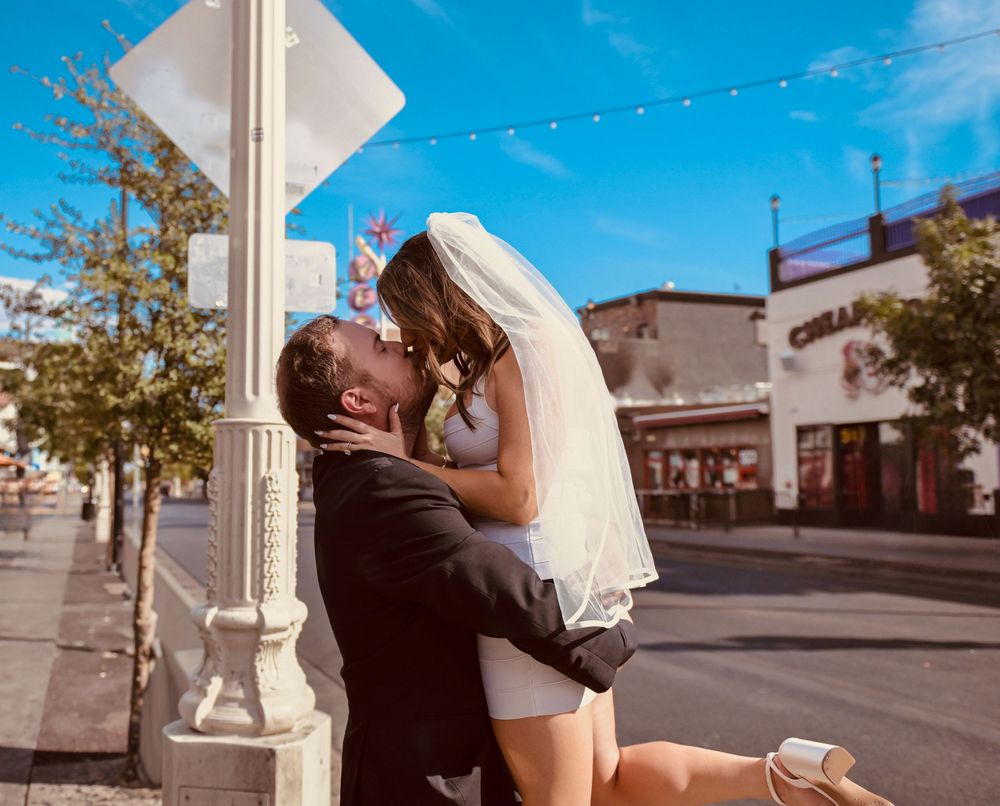 Newlyweds kissing passionately on a sunny street corner, bride in white dress and veil, groom in black suit.