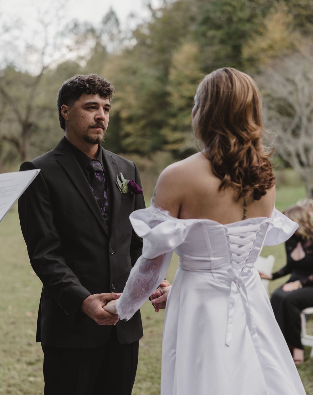 Groom in a dark suit holds hands with a bride in a white off-shoulder lace-up gown during an outdoor wedding ceremony.