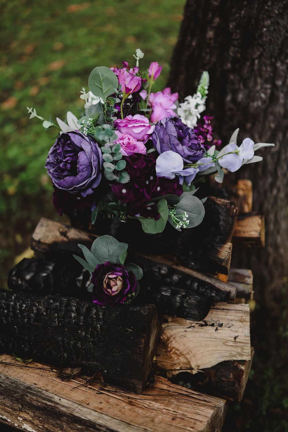 Purple and white floral bouquet with eucalyptus leaves resting on stacked charred and natural firewood beside a tree trunk.
