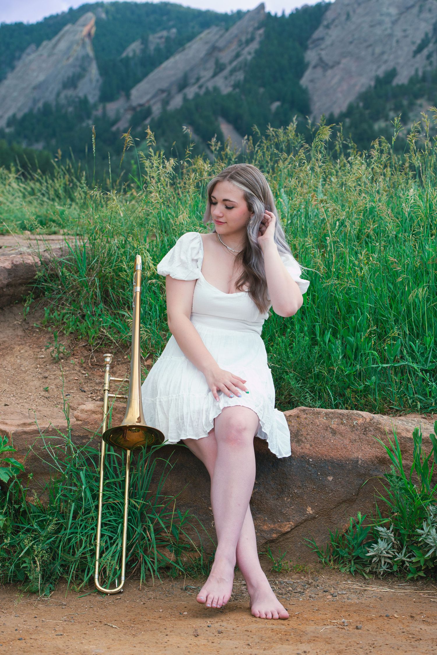 Young woman in white dress sits on rock with trombone beside her, Flatirons mountains in background, Boulder CO.