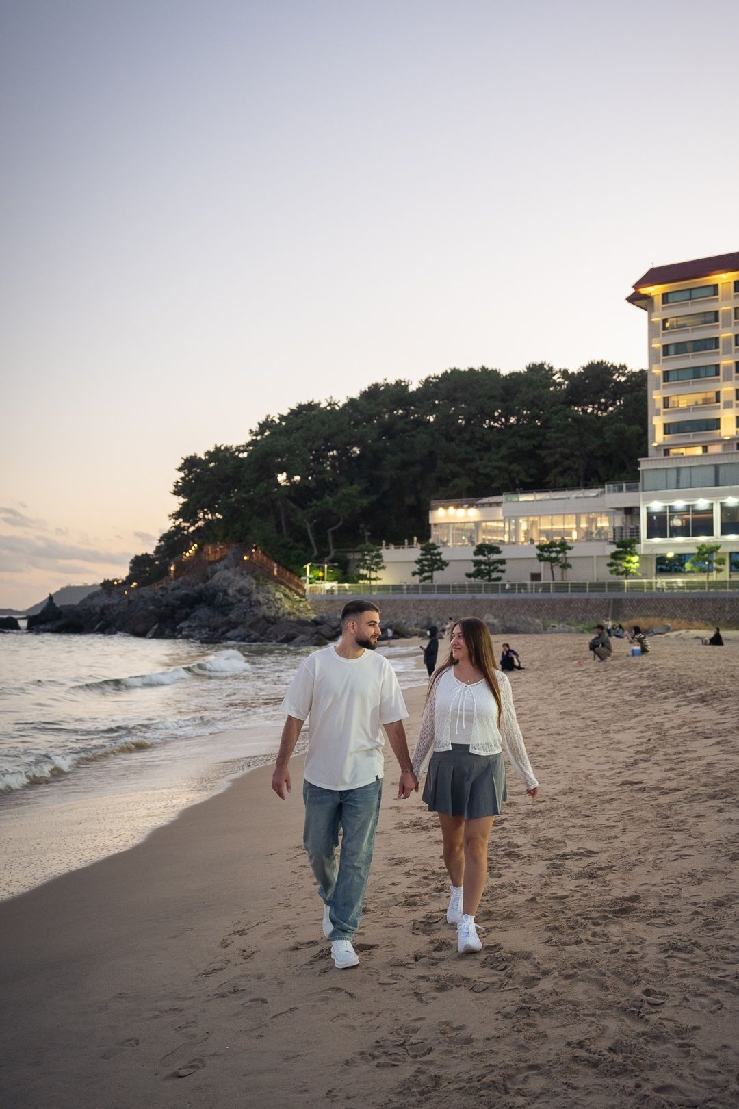Couple holding hands walking on sandy beach at sunset with hotel and trees in background.