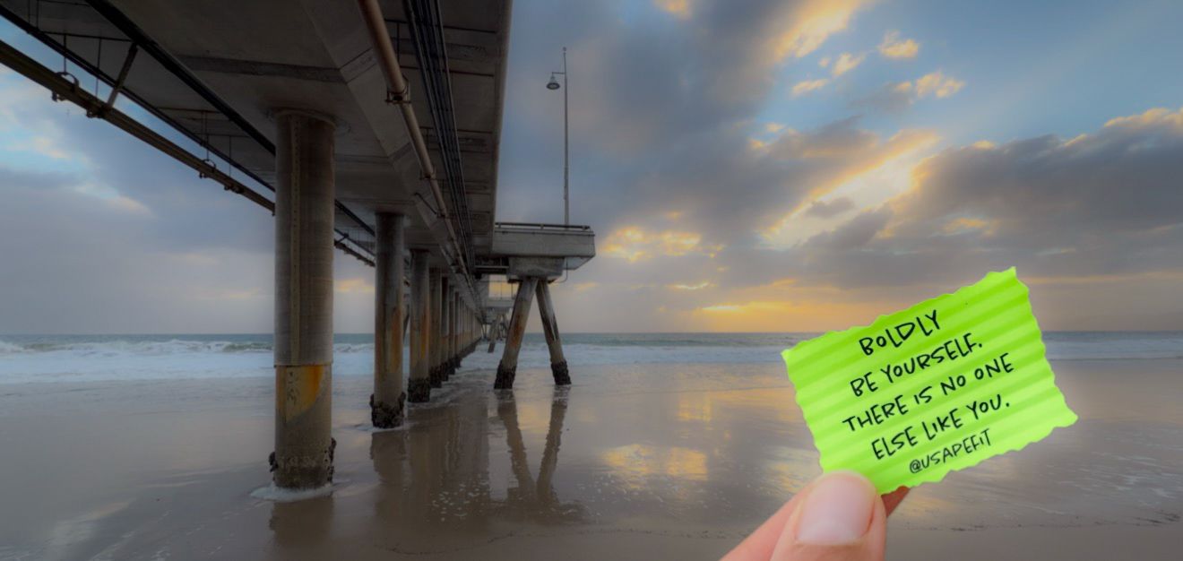 Hand holding a green sticky note with inspirational quote on a beach pier at sunset with dramatic cloudy sky.