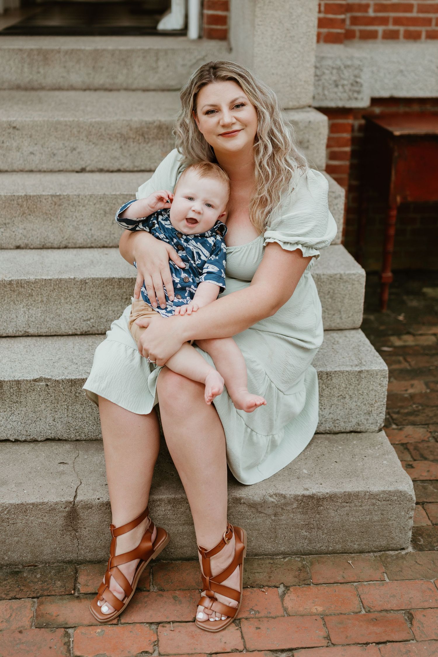 Mother and baby sit together on brick steps in summer dress and sandals.