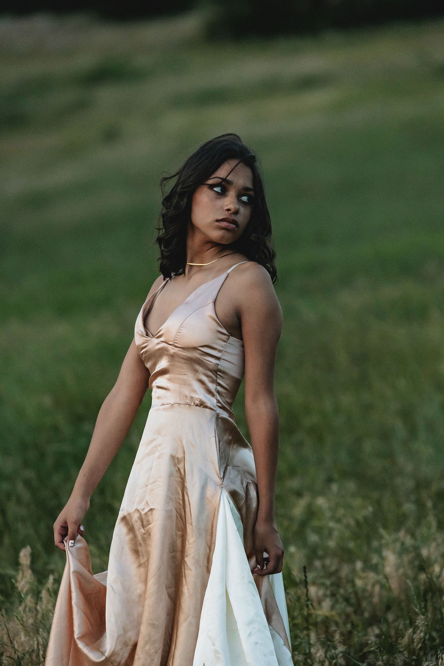 A young woman in a satin champagne slip dress stands gracefully in a lush green field.