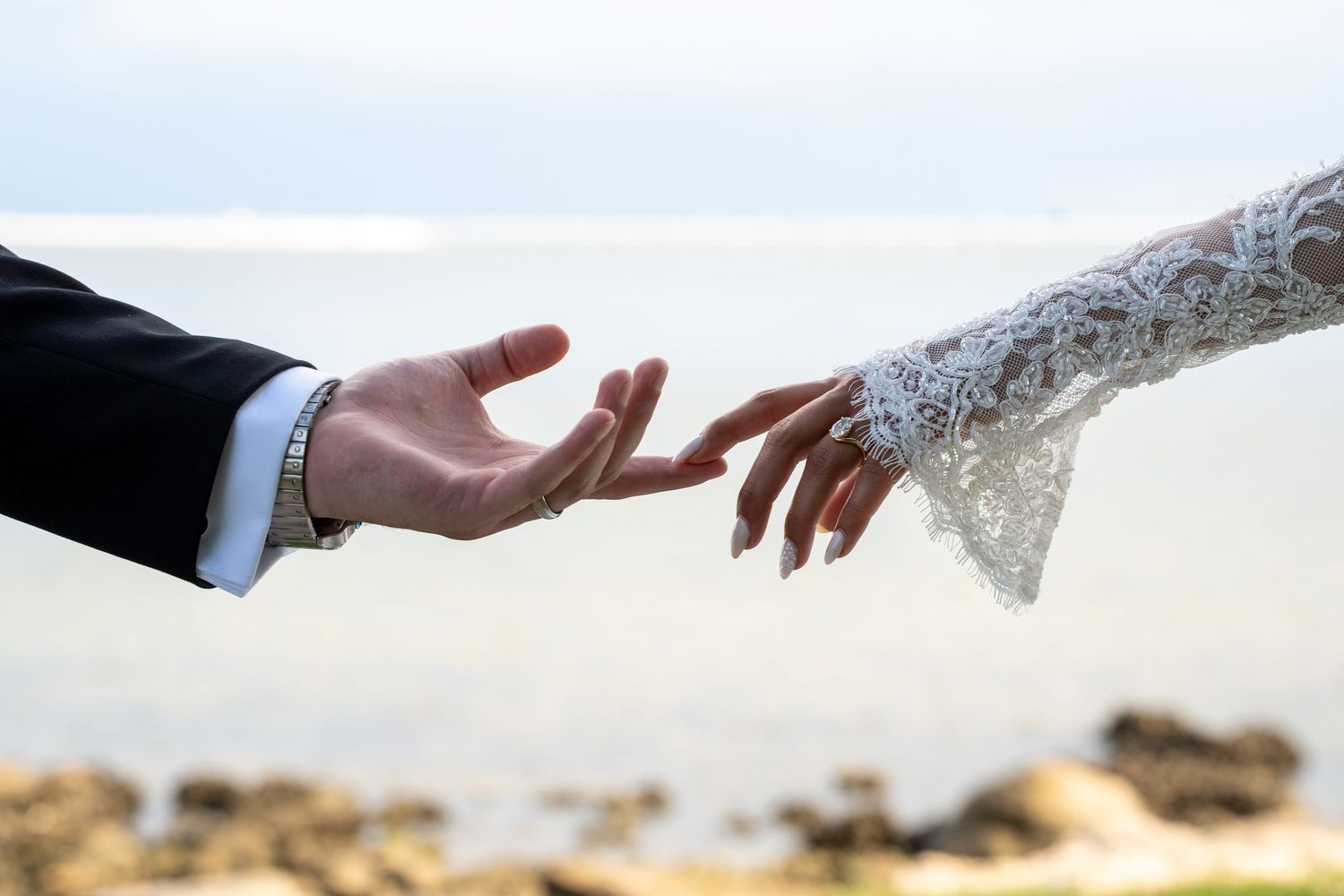 Bride and groom reaching hands toward each other near ocean shore, wedding lace sleeve visible.