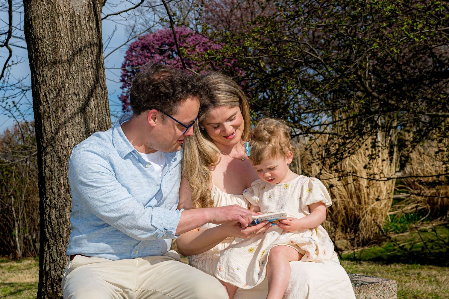 A young family sits outdoors under a tree, parents smiling while showing their toddler daughter something on a small device.