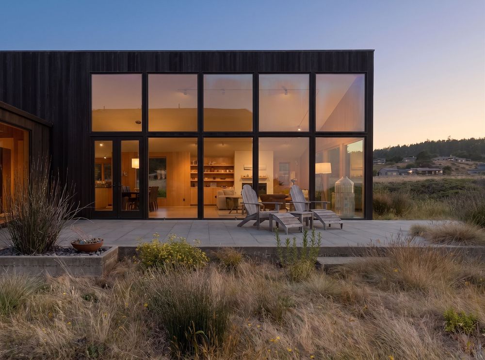 Modern black wood home with floor-to-ceiling windows illuminated at dusk, surrounded by natural grasses and rolling hills.