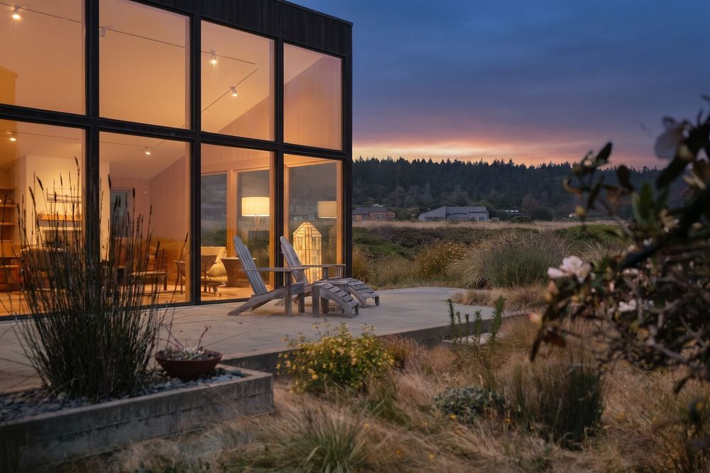 Modern glass house with warm interior lights glowing at dusk, surrounded by natural grasses with a scenic sunset sky.