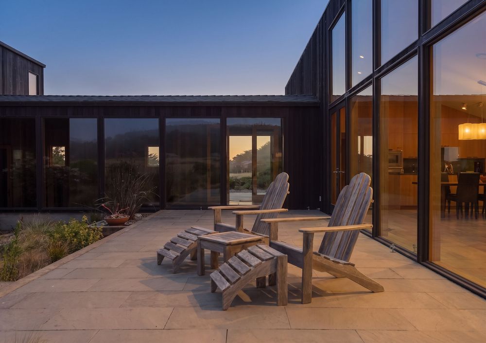 Modern black home courtyard with wooden Adirondack chairs on stone patio at dusk, featuring floor-to-ceiling windows.