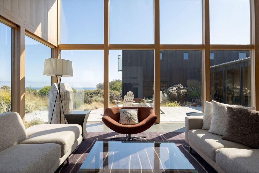 Modern living room with floor-to-ceiling windows, neutral sofas, leather chair, and panoramic coastal views.