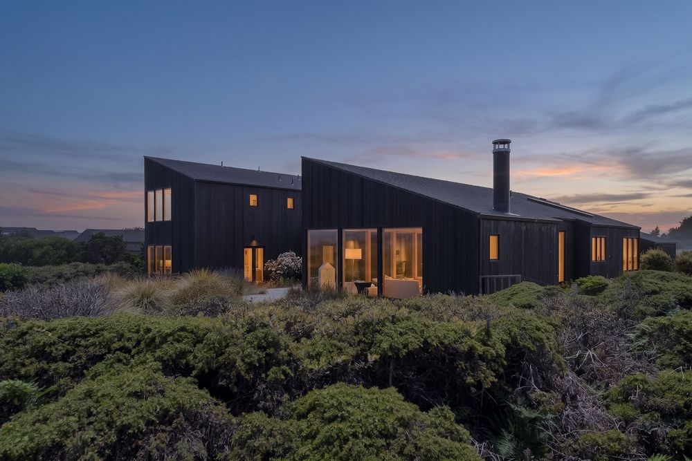 Dark-clad modern house surrounded by coastal shrubs at dusk, with warm interior lights glowing through large windows.