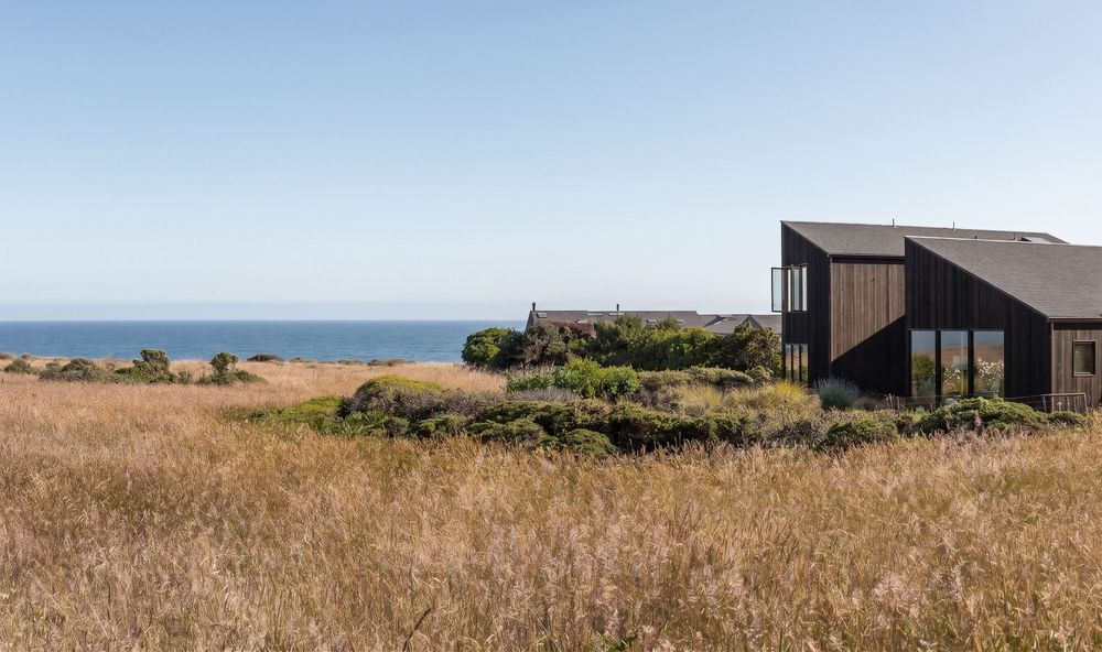 Dark wood coastal home surrounded by golden grasses with ocean views under a clear blue sky.