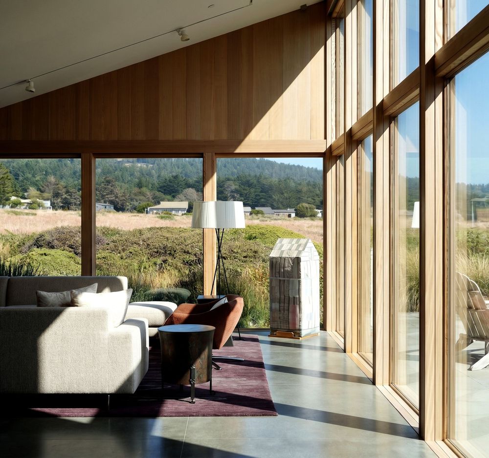 Modern living room with floor-to-ceiling wood-framed windows overlooking a scenic rural landscape with rolling hills.
