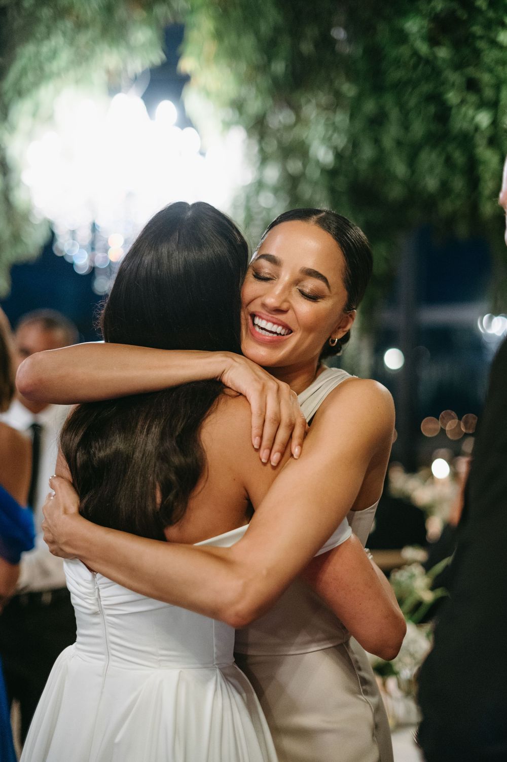 Two people in white dresses share emotional embraces during an outdoor evening celebration.