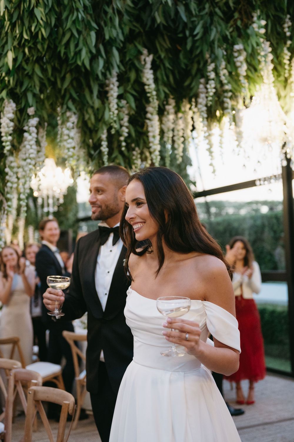 Wedding guests mingle and celebrate during an outdoor reception under hanging floral decorations at dusk.