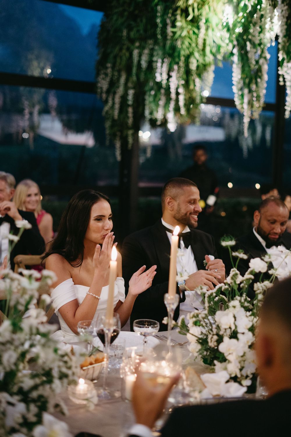 Wedding guests enjoy an elegant outdoor reception under hanging greenery and white floral centerpieces at night.