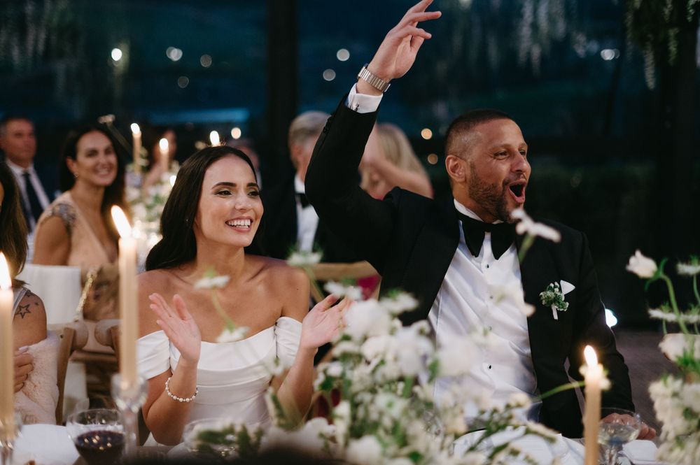 Wedding guests raise their glasses in celebration during an outdoor reception under string lights and trees.
