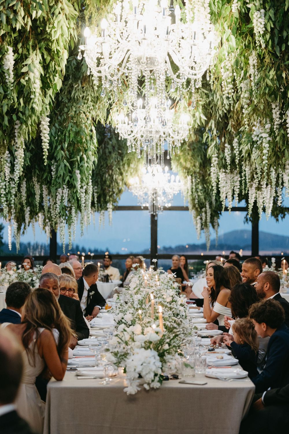 Elegant wedding reception under cascading white wisteria with long tables and guests seated beneath floral canopy.