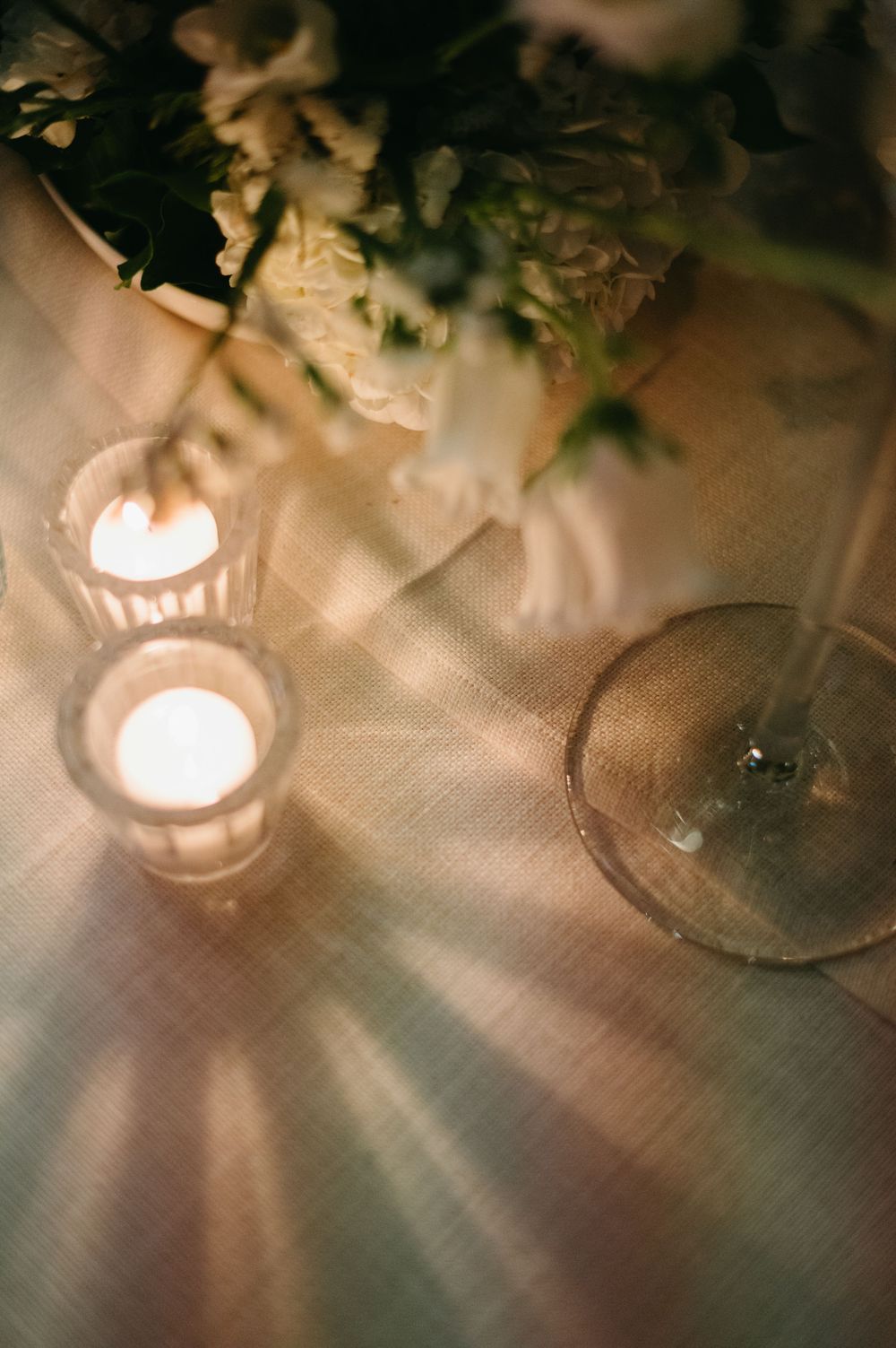 Soft glowing candlelight creates a romantic ambiance on a textured tablecloth with delicate greenery accents.
