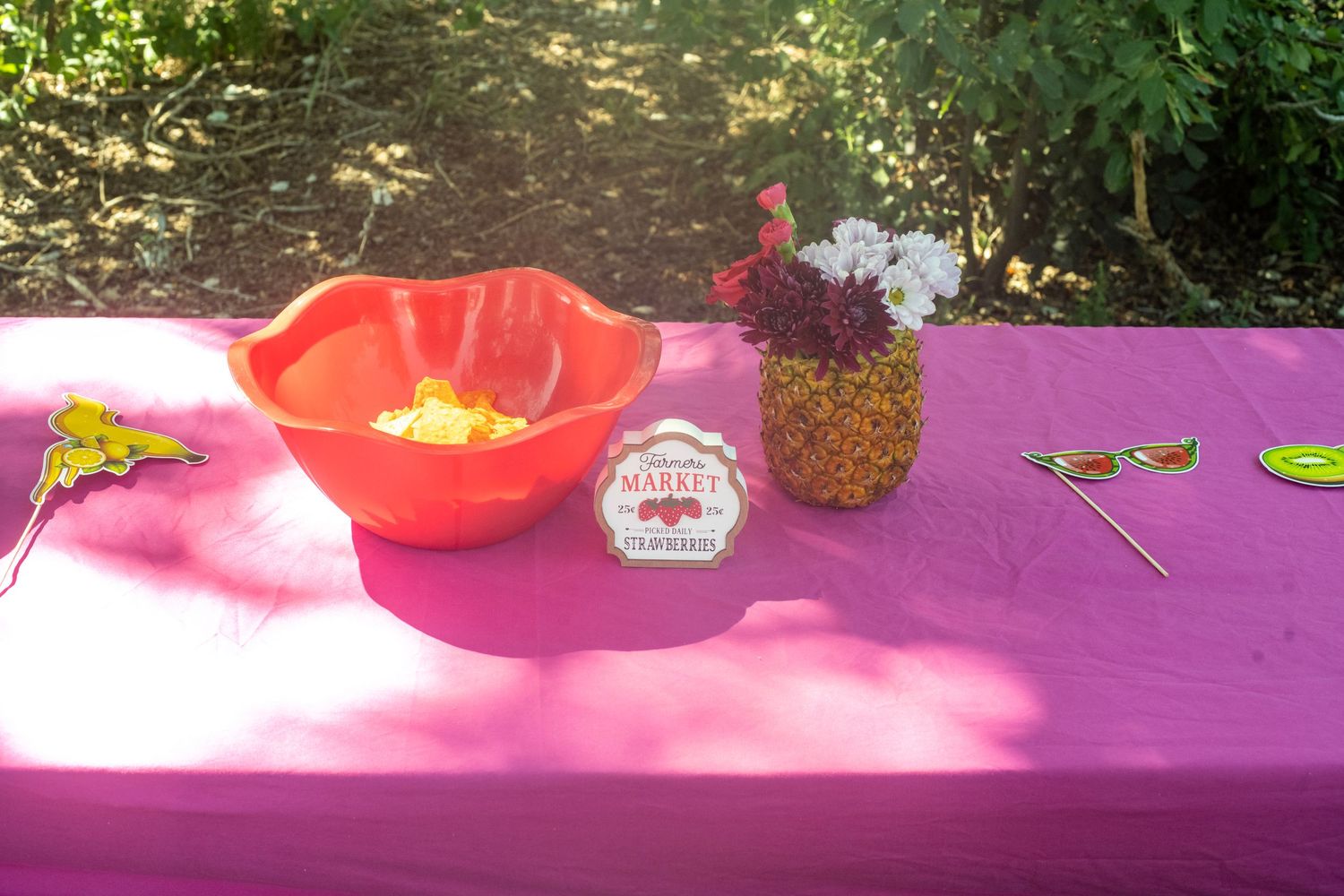 Pink tablecloth outdoor party setup with orange bowl, pineapple vase with flowers, and colorful party props in garden.