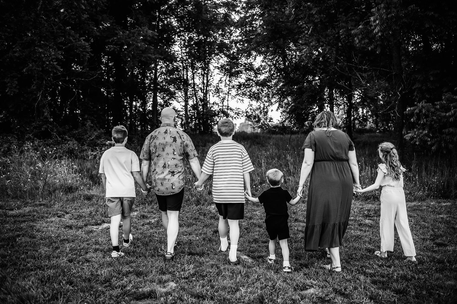 Black and white photo of a family holding hands walking through a grassy field toward trees at sunset.