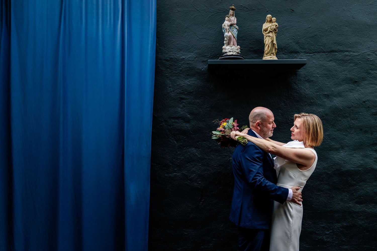 Elegant couple embracing near blue curtain and dark wall with figurines on shelf during wedding ceremony.