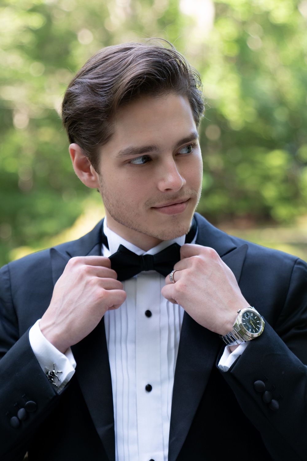Elegant groom in black tuxedo adjusting bow tie outdoors with green trees in background.