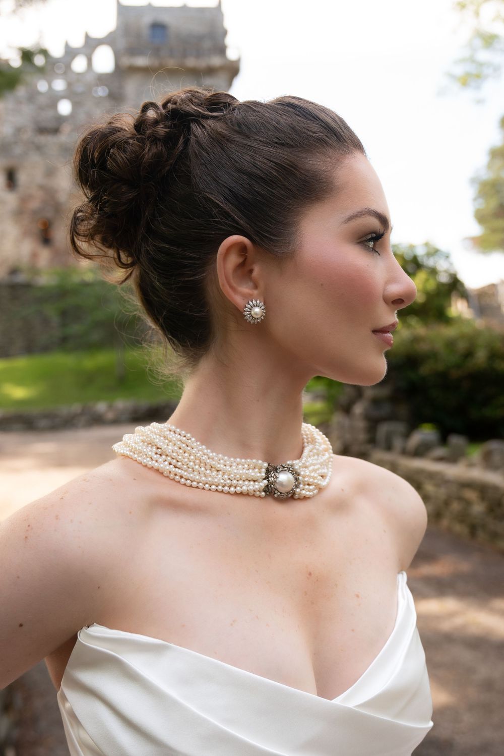 Bride in profile wearing pearl choker necklace and updo hairstyle with castle ruins backdrop.