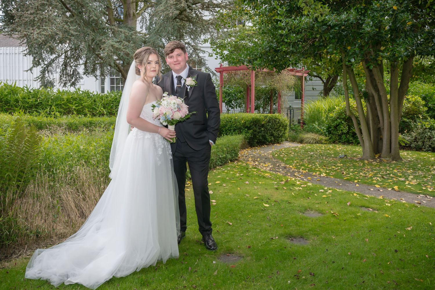 Bride in white gown and veil holds bouquet beside groom in black suit, posing in lush garden setting.