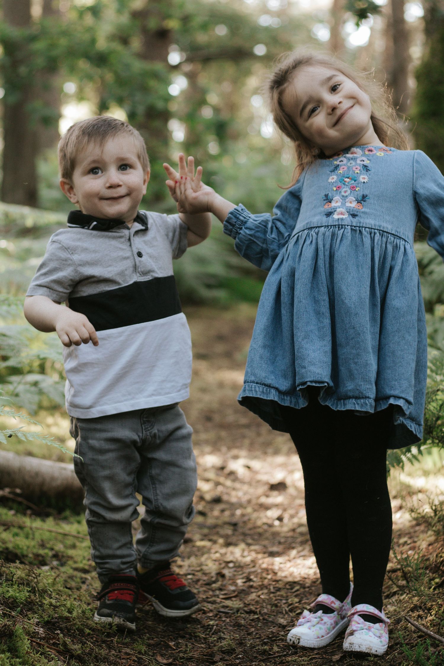 Two young children high-fiving in a forest, a boy in gray and a girl in a blue dress with embroidery.