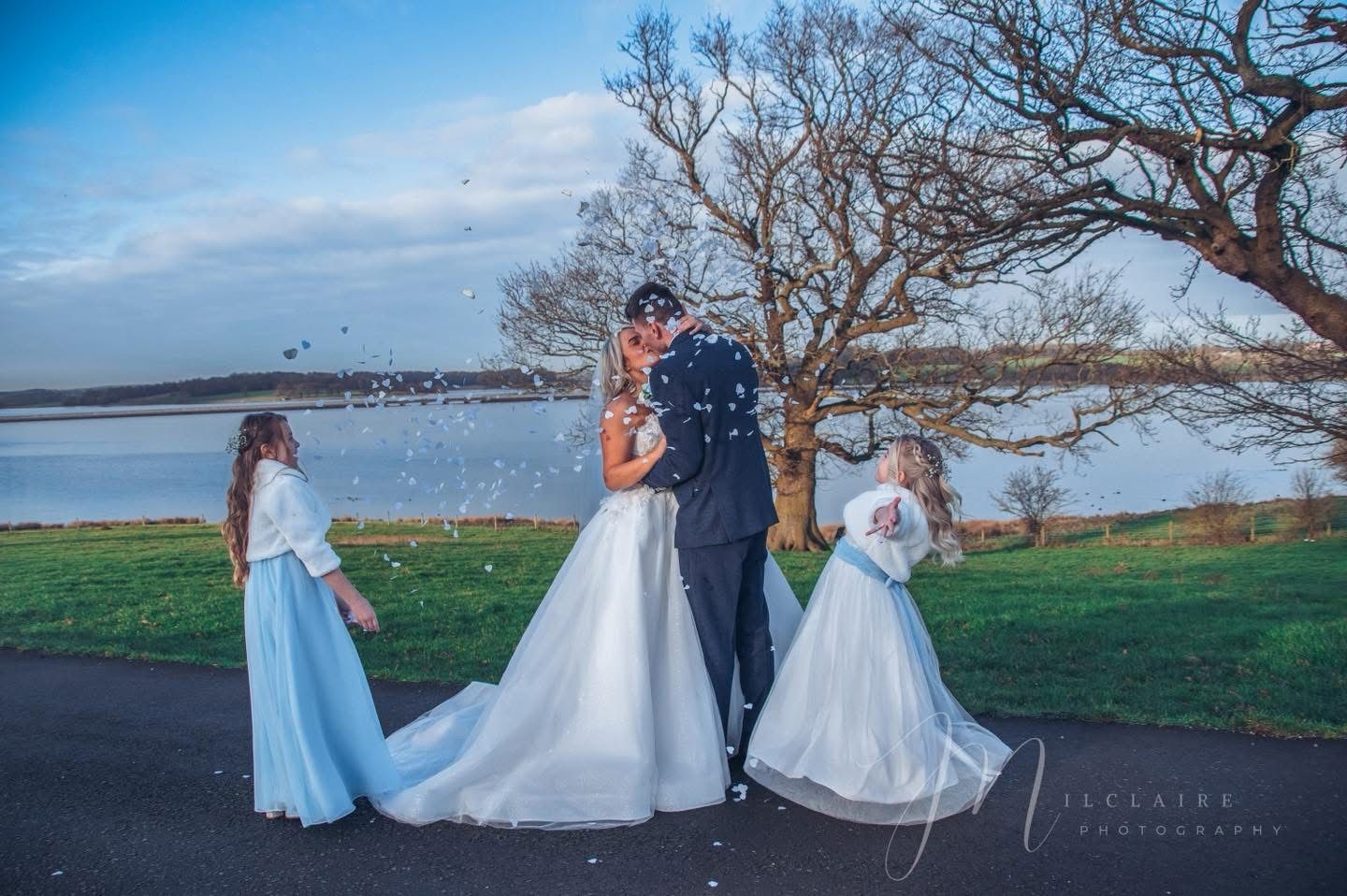 Bride and groom kissing while two flower girls in blue dresses throw confetti by a lakeside. Rugeley