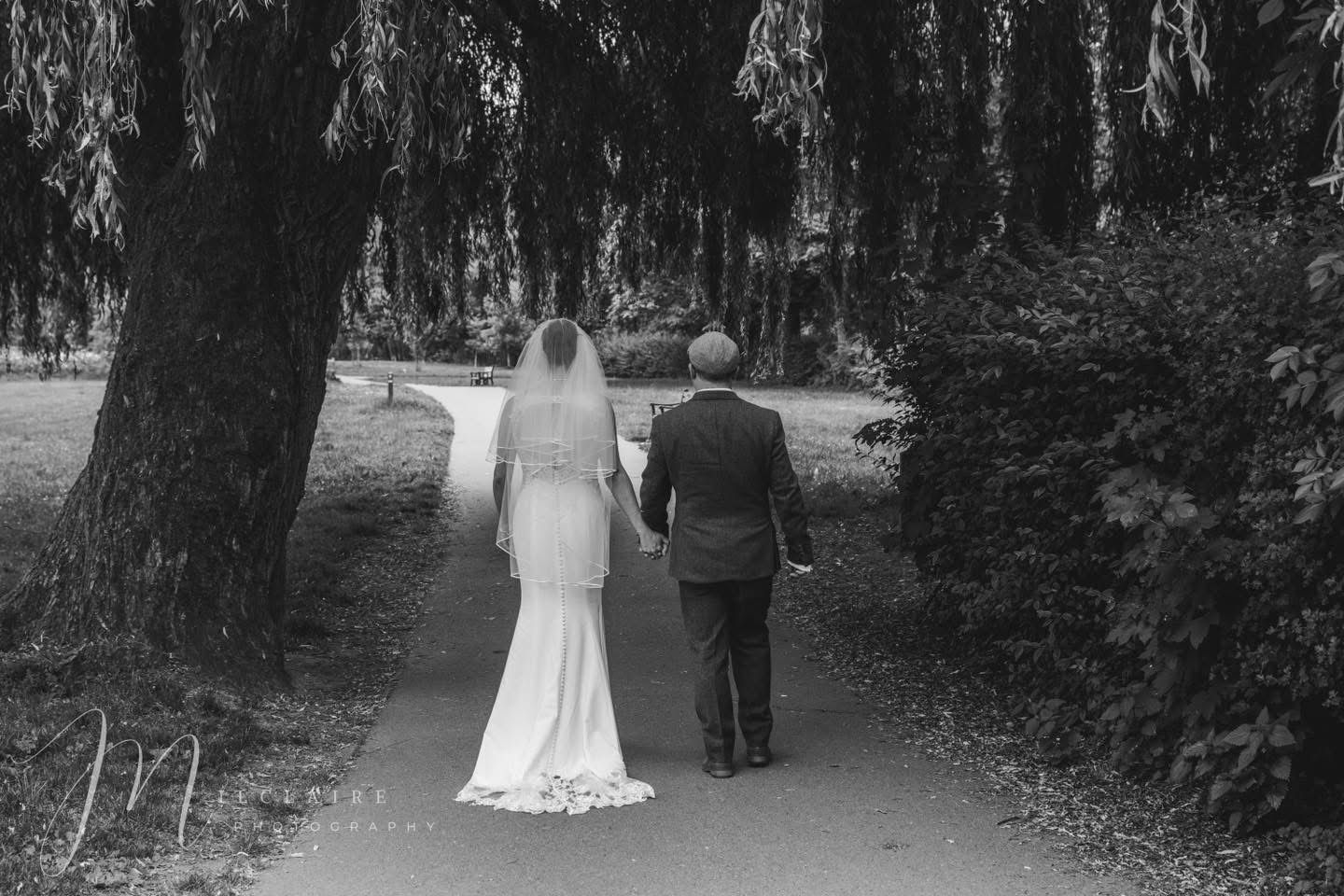 Black and white photo of a bride and her escort walking hand in hand down a tree-lined garden path. Coventry